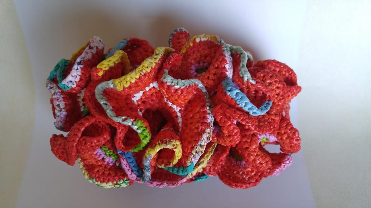 A dense cluster of ruffled, crocheted coral-like forms in bright red, their frilled edges trimmed in yellow, turquoise, green and pink.