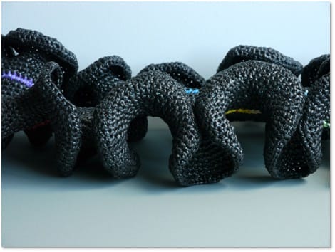 A black crocheted form folded into regular deep waves, flecked here and there with purple, yellow and blue.