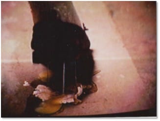 A dark blurred image of a black form standing over a small pale spill on a pink-grey surface.