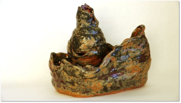 A tall, rounded, dark mottled form rising from a wide craggy bowl, its surface flecked with rust, purple and brown.