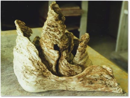 A pale clay sculpture of jagged upright spires streaked with brown, rising from a smooth curved base on a workshop table.