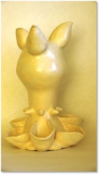 A pale yellow glazed sculpture with two pointed ear-like tips above a bulbous body, seated in a ring of open petal shapes.