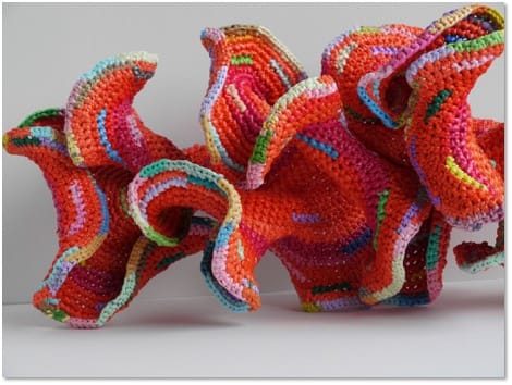 An undulating orange crocheted form with deeply rippled frilled edges picked out in pink, green and blue.